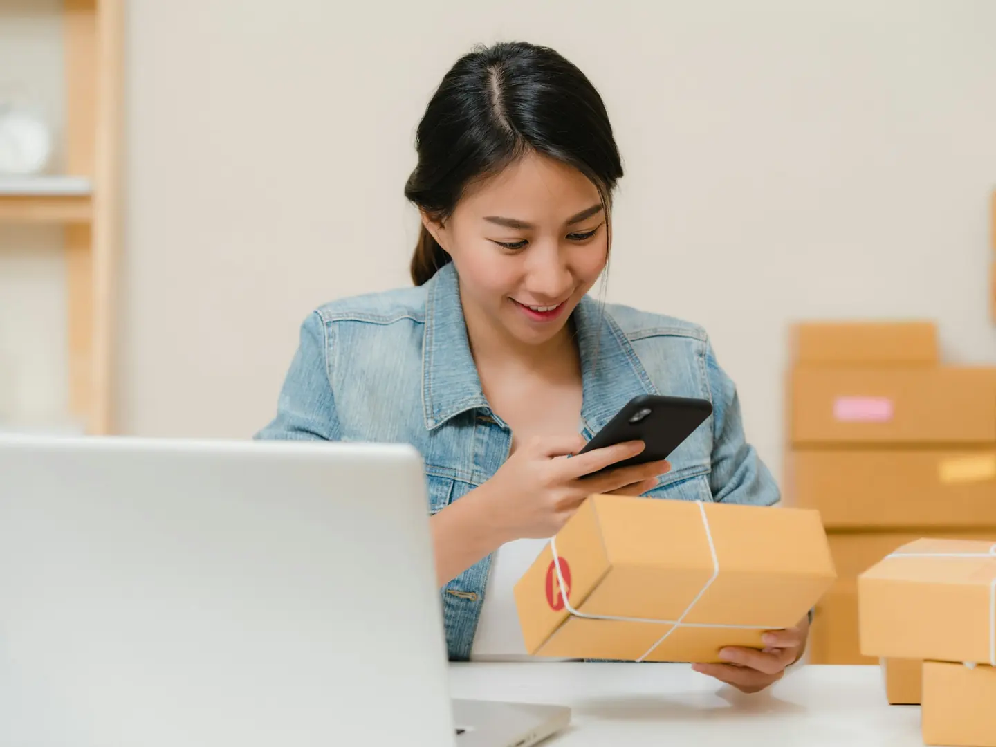 An online store owner checks an order on her phone next to her laptop and packed parcels.